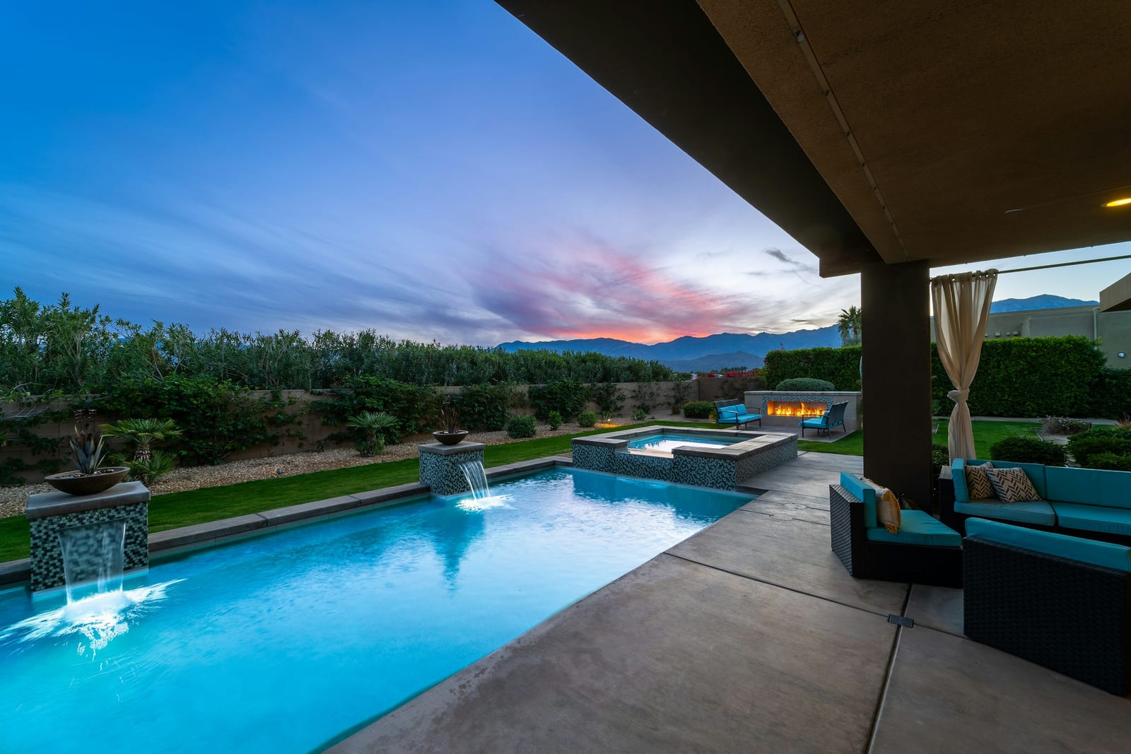 A raised spa spilling into a pool past glass-tile scupper bowls, with desert mountains turning pink at sunset.