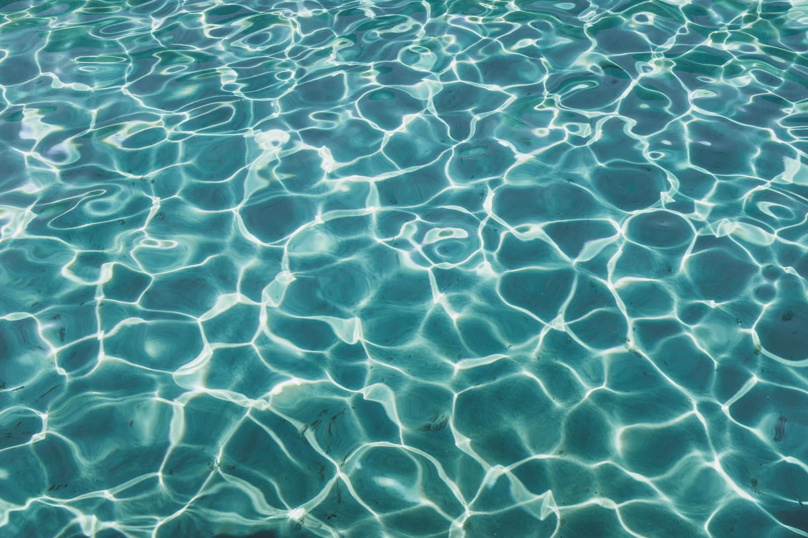 Sunlight caustics rippling across clear turquoise pool water over a light plaster finish.
