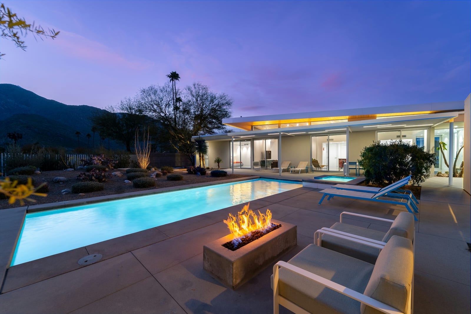 A desert-modern home at twilight with a linear fire pit, a lit lap pool and spa, ocotillo and a mountain silhouette behind.